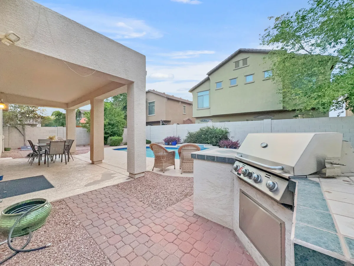 A sun-drenched backyard scene featuring a sparkling swimming pool, a fully equipped outdoor grill and kitchen area, a dining table perfect for gatherings, and comfortable seating. The space is set up for a range of outdoor activities, from swimming and barbecuing to relaxing and socializing.