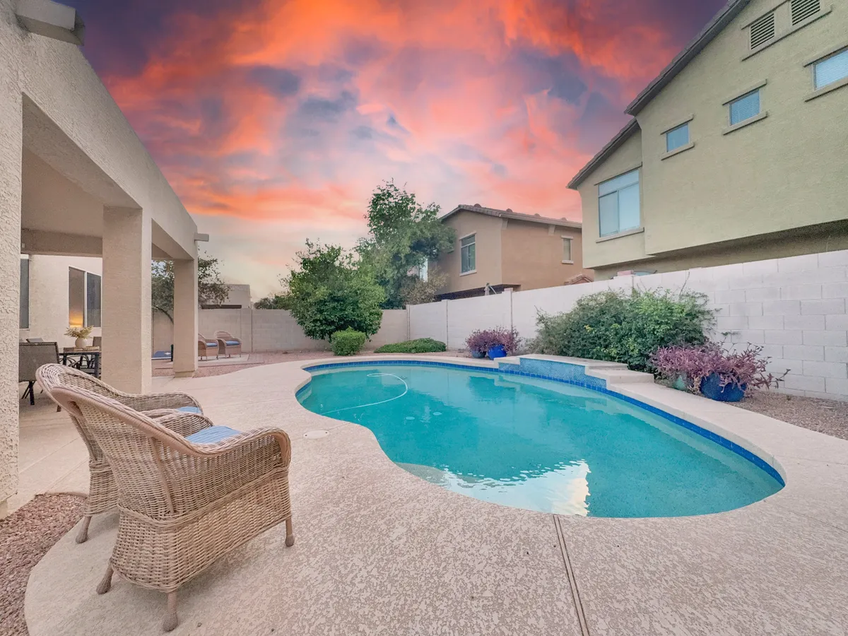 A tranquil exterior scene featuring a sparkling swimming pool surrounded by wicker chairs, all set against the backdrop of a colorful sunset sky. The inviting setup suggests a space perfect for swimming, lounging, and social gatherings at dusk.