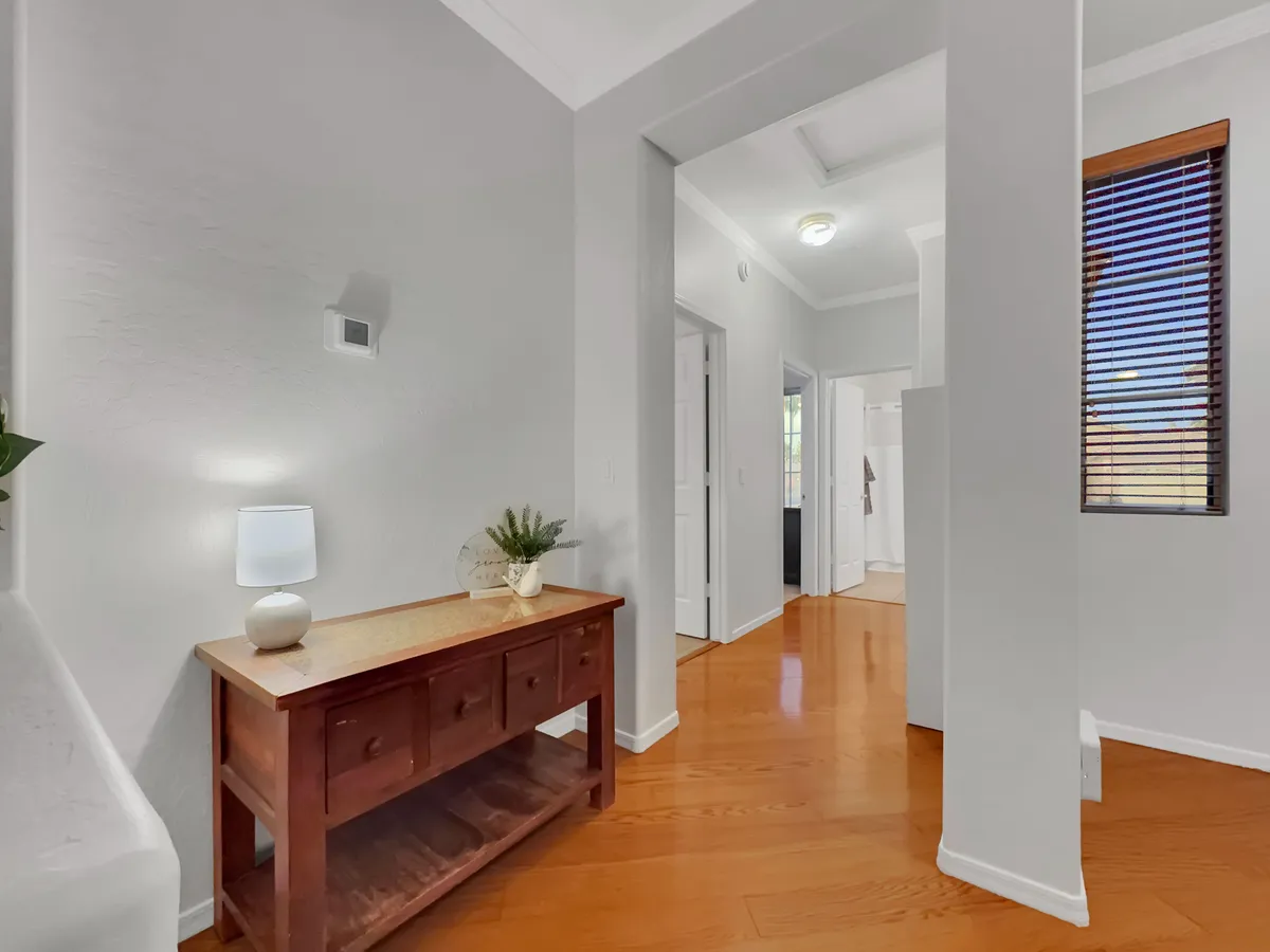 A wooden console table topped with a decorative plant and lamp sits beside a sunlit window with blinds. The setup is positioned in what appears to be an entryway or transitional area, reinforcing a sense of welcome and daily comings and goings.